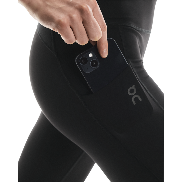En kvinde i sorte On Performance Tights Winter, Dame, lægger en sort smartphone i sidelommen. Strømpebukserne har et lille On-logo nær lommen og er ideelle til vinterbrug.
