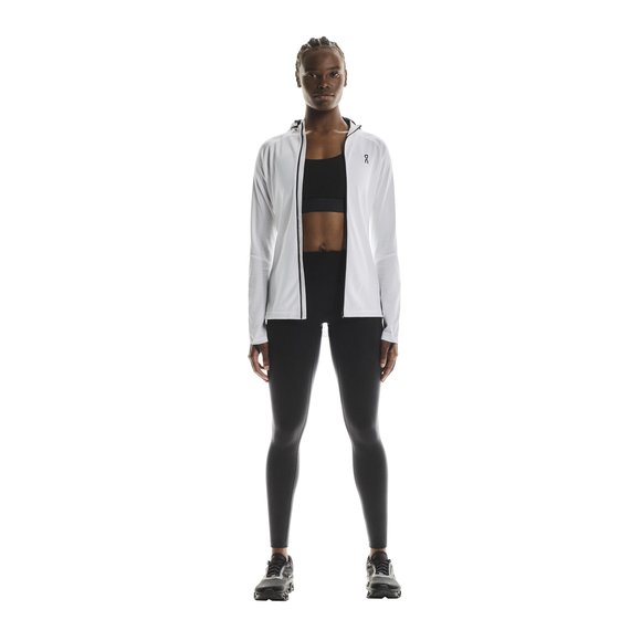 En kvinde kigger fremad mod en almindelig hvid baggrund iført On Performance Tights, Dame, Black med en hvid jakke med lynlås over en sort sports-bh og sorte sportssko.