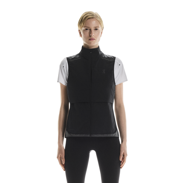 En kvinde ser frem for sig iført On Weather Vest til kvinder i Black/Eclipse over en hvid kortærmet skjorte og sorte leggings på en almindelig hvid baggrund.