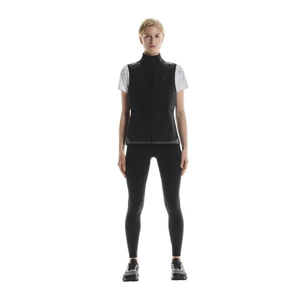 En person kigger fremad iført On Weather Vest til kvinder i Black/Eclipse over en hvid kortærmet skjorte, sammen med sorte leggings og sorte sportssko på en almindelig hvid baggrund.