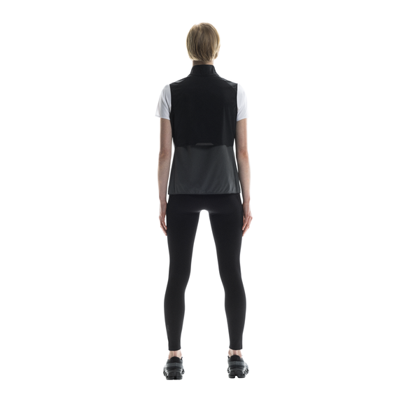 En person står med ansigtet vendt mod kameraet iført On Weather Vest, Dame, Black/Eclipse over en hvid skjorte med sorte leggings og sorte sko på en almindelig hvid baggrund.