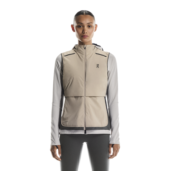 En kvinde kigger fremad, iført On Weather Vest Dame i Desert/Eclipse over en lysegrå langærmet skjorte og sorte leggings, klar til vejrbeskyttelse, der løber mod en almindelig hvid baggrund.