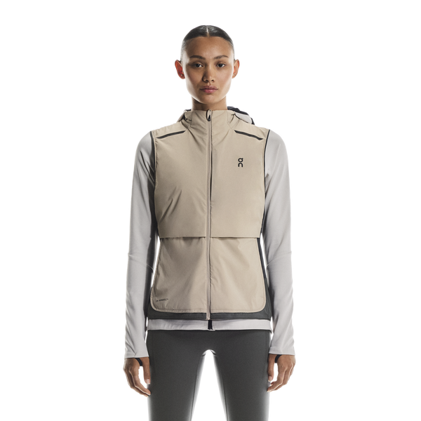 En kvinde kigger fremad, iført On Weather Vest Dame i Desert/Eclipse over en lysegrå langærmet skjorte og sorte leggings, klar til vejrbeskyttelse, der løber mod en almindelig hvid baggrund.