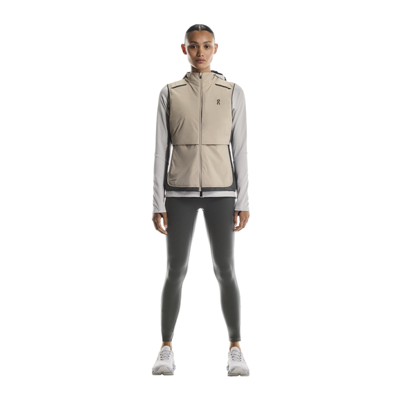 En kvinde ser frem for sig iført en On Weather Vest Dame i Desert/Eclipse over en lysegrå langærmet top, mørkegrå leggings og hvide sneakers - perfekt vejrbeskyttelse til løb - på en almindelig hvid baggrund.