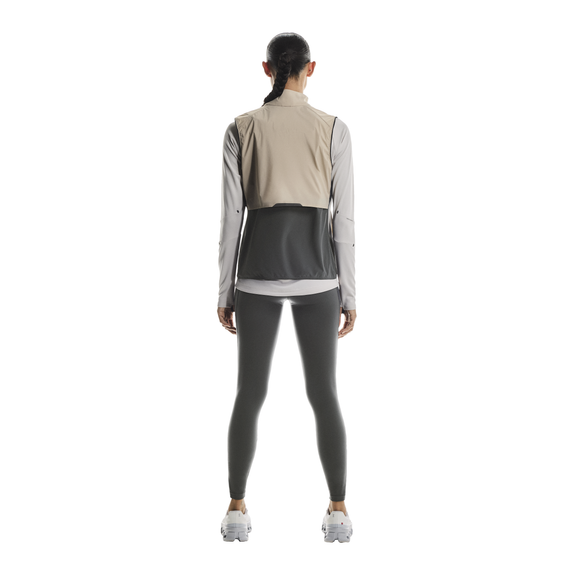 En kvinde kigger væk fra kameraet iført On Weather Vest Dame i Desert/Eclipse - perfekt til beskyttelse mod vejret på løbeturen - over en grå langærmet skjorte, mørkegrå leggings og hvide sneakers på en almindelig hvid baggrund.