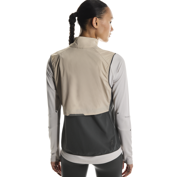 En kvinde med mørkt flettet hår ses bagfra iført On Weather Vest, Dame, i Desert/Eclipse (beige og mørkegrå) over en lys langærmet skjorte - perfekt vejrbeskyttelse løb.