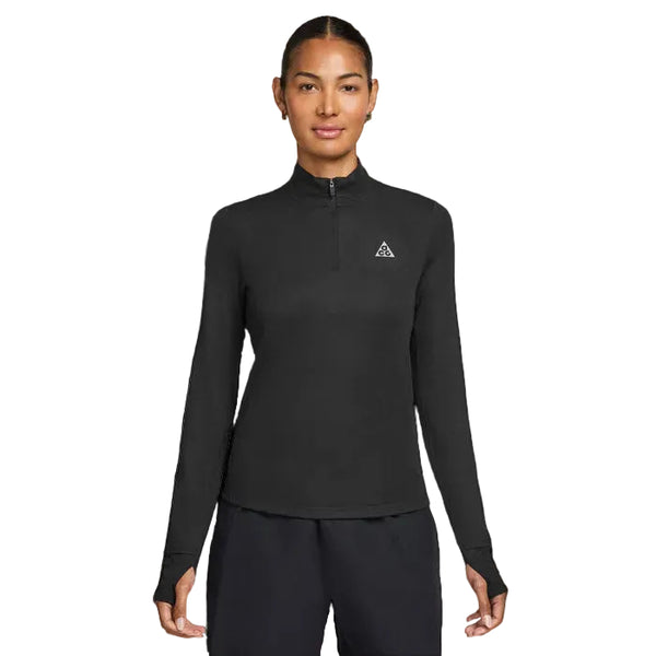 En kvinde iført en sort Nike ACG Half Zip Running Top, Dame, Black/Black/Summit White med huller til tommelfingrene og et lille trekantet logo på venstre bryst, står med front mod en hvid baggrund.