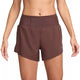 En kvinde iført Nike Dri-FIT Trail Mid-Rise 4" Brief-Lined Running Shorts i Red Sepia/Black, med elastisk linning og afslappet pasform, står med armene lidt ud til siderne mod en ensfarvet baggrund.