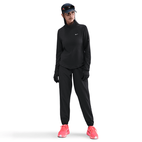 En person står på en hvid baggrund iført helt sort Nike dame løbetøj med Therma-FIT Turtle-Neck Running Top i Black/Reflective Silv sammen med handsker, visir, solbriller og lyserøde sportssko.