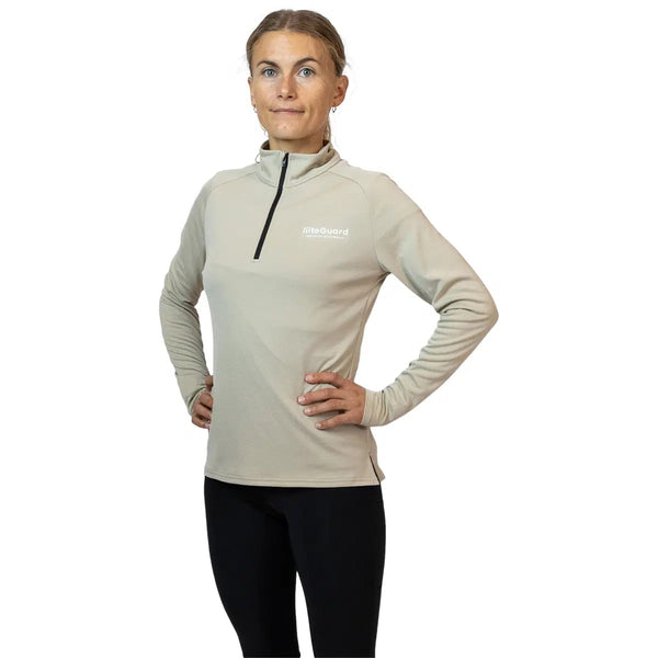 En person står med hænderne på hofterne, iført en beige Liiteguard Ground-Tech 1/4 Zip, Dame med RiteGuard-logo og sorte leggings, og kigger fremad mod en almindelig hvid baggrund.
