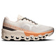 On Cloudmonster 2 Dame Ivory/Tangerine har en tyk geometrisk sål med store udskæringer, responsivt Helion-superfoam, en overdel i beige og orange mesh, sorte og hvide snørebånd og "ON RUNNING" langs siden.