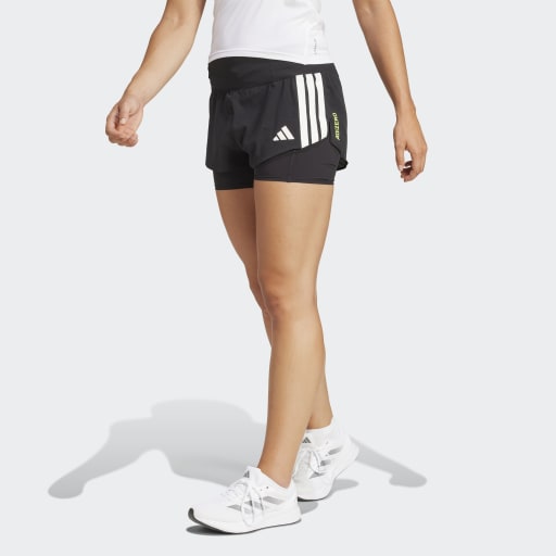 En person er iført en hvid skjorte, Adidas Adizero 2I1 Shorts, Dame i sort med hvide striber, og hvide sneakers på en almindelig lys baggrund. Kun den nederste halvdel er synlig. Ideel til løb med behageligt kompressionsmateriale.