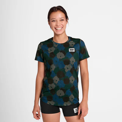 En person står på en almindelig lys baggrund, smiler og er iført Saysky W Flower Combat T-Shirt, Dame, Blue Aop med sorte shorts. Både t-shirten og shortsene har små rektangulære lapper.