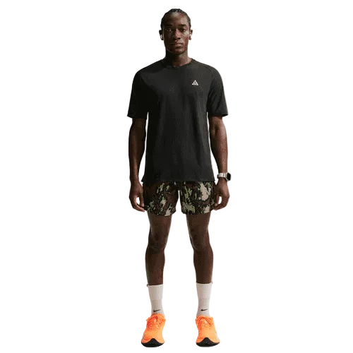 En mand ser frem for sig i en Nike ACG "Solar Chase" Trail Running Top (Black/Black/Summit White), camouflageshorts, hvide lægsokker, lyseorange sportssko og et ur på venstre håndled med et neutralt ansigtsudtryk.