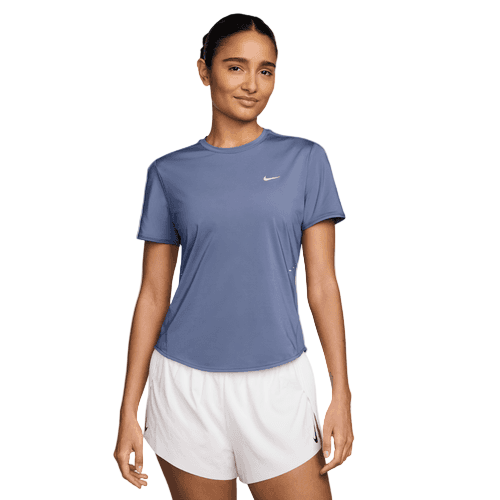 En person står med ansigtet fremad iført en Nike Swift SS Løbetop til kvinder i World Indigo med reflekterende sølvdetaljer og hvide sportsshorts. Baggrunden er almindelig hvid.