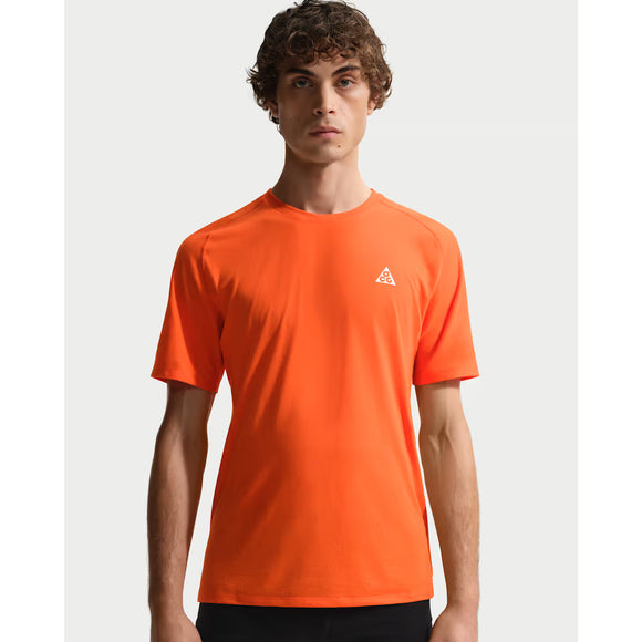 En ung mand med krøllet brunt hår bærer en lysende orange Nike ACG Dfadv Solar Chase Ss træningstop herre (Safety Orange/Summit White) med et lille hvidt trekantet logo på venstre bryst, der står på en almindelig lysegrå baggrund.