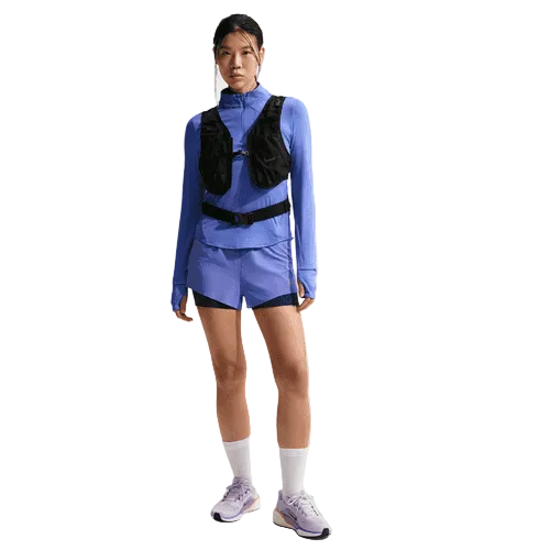 En person står og kigger fremad iført en Nike Swift UV Half Zip Running Top til kvinder i Sapphire/Reflective Silver med matchende shorts, en sort vest, hvide strømper og lyse sportssko på en almindelig hvid baggrund.