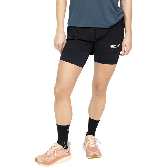 En person iført blå skjorte, sorte Liiteguard Glu-Tech Running 2In1 Shorts (Dame) med "Queen" på benet, sorte sokker og lyserøde løbesko står på en almindelig hvid baggrund. Vedkommendes hoved er ikke synligt på billedet.