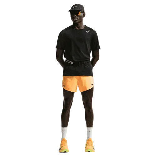 En person står på en hvid baggrund iført en sort Nike-kasket, sort t-shirt, AeroSwift 4In Brief-Lined Running Shorts by Nike i Laser Orange/Black, hvide sokker, gule Nike-sko og solbriller. Hænderne er bag ryggen.