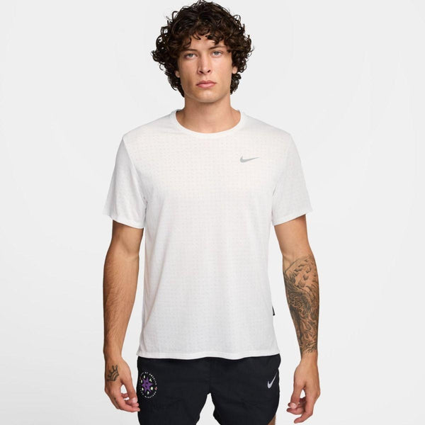 En mand med krøllet hår er iført en hvid Nike Miler kortærmet løbetop, Herre (Summit White/Reflective Silver) og sorte Nike shorts. Han står med ansigtet fremad mod en almindelig hvid baggrund med et neutralt udtryk.