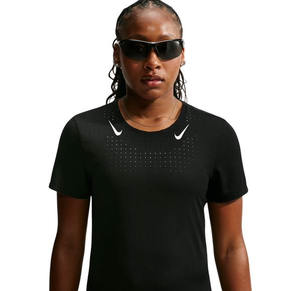 En person med flettet hår står med ansigtet vendt fremad foran en ensfarvet hvid baggrund og har en sort Dri-FIT ADV Running Top, Dame fra Nike, på. Trøjen har et hvidt Nike-logo på brystet og er designet som svedtransporterende løbetøj. Personen bærer desuden mørke solbriller.