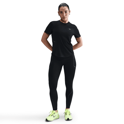 En kvinde iført en sort kortærmet sportstrøje, Nike Therma-FIT ADV High-Waisted 7/8 Reflective Running Leggings i Black/Dk Smoke Grey og neongule løbesko står med ansigtet fremad og hænderne bag ryggen mod en hvid baggrund.
