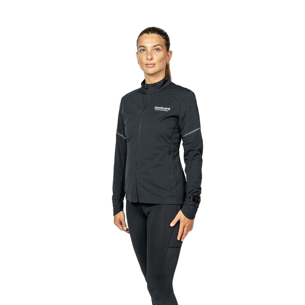 En kvinde står med front mod en hvid baggrund iført Liiteguards Hydro-Tech Running Jacket, Dame, Black og sorte leggings. Jakken har et lille logo på brystet, håndledsdetaljer på venstre ærme og leverer en elegant, atletisk stil.