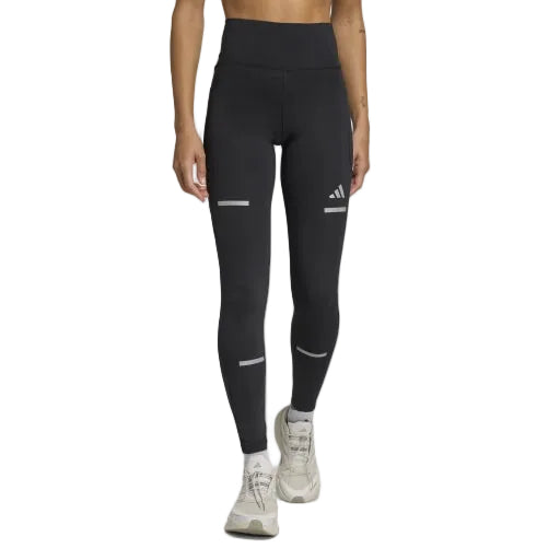 En kvinde iført sorte Adi365 Leggins fra Adidas med reflekterende detaljer og et lille logo på venstre lår, parret med lyse sportssko, står på en ensfarvet lysegrå baggrund.
