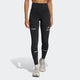 En kvinde iført sorte Adi365 Leggins fra Adidas med reflekterende detaljer og et lille logo på venstre lår, parret med lyse sportssko, står på en ensfarvet lysegrå baggrund.