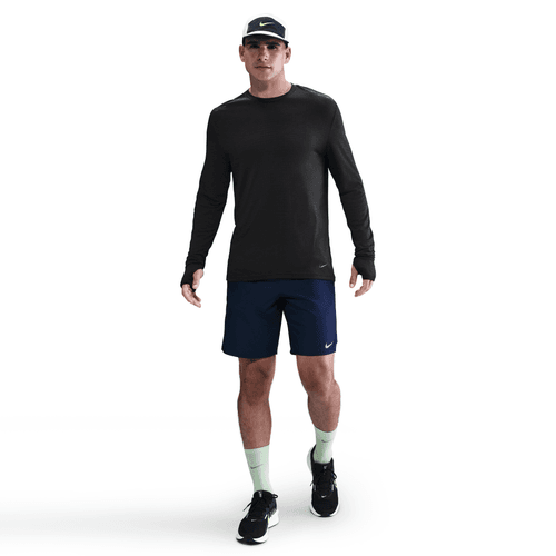 En mand iført en Nike Dri-FIT ADV Wool langærmet løbetop i sort/antrazit, marineblå sportsshorts, lysegrønne sokker, sorte løbesko og en mørk kasket går frem mod en almindelig hvid baggrund.