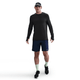 En mand iført en Nike Dri-FIT ADV Wool langærmet løbetop i sort/antrazit, marineblå sportsshorts, lysegrønne sokker, sorte løbesko og en mørk kasket går frem mod en almindelig hvid baggrund.