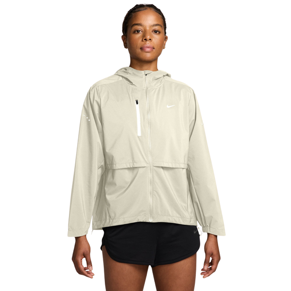 En person iført en lysbeige Nike Storm-FIT-løbejakke med hætte, Dame, i farverne Sail/Reflective Silv samt sorte sportsshorts.
