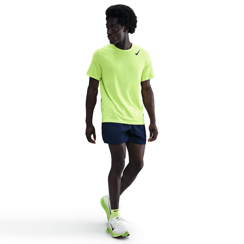 En person står på en almindelig hvid baggrund iført Nike Dri-FIT ADV Short-Sleeve Running Top til mænd i Volt/Black, parret med marineblå løbeshorts og hvide løbesko med neongule detaljer.