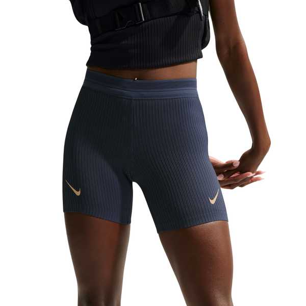 En person iført Nike AeroSwift Tight løbeshorts til kvinder i farvekombinationen Thunder Blue/Black/White/Linen og en sort, ærmeløs top står med armene bag ryggen foran en hvid baggrund. Det svedtransporterende design sikrer komfort under alle former for aktivitet.
