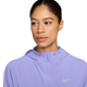 En kvinde har Nike Swift Repel Packable Running Jacket på i farven Purple Pulse/Reflective Silver med hætten nede og kigger let til siden. Dette sporty stykke tøj, der er inspireret af sammenpakkelige løbejakker, er perfekt til aktive dage. Baggrunden er ensfarvet hvid.