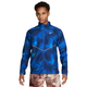 En person iført en Nike Miler UV Repel-løbejakke til mænd i farven Midnight Navy/Reflective Silver med et hvidt logo, kombineret med mønstrede shorts, står med ansigtet vendt fremad foran en ensfarvet hvid baggrund.