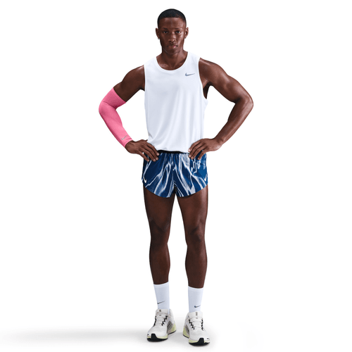 En mand står med ansigtet fremad og hænderne på hofterne, iført en hvid tanktop, Nike Aeroswift 2" Shorts til mænd i Coastal Blue/White, hvide sokker, sportssko og et lyserødt ærme på højre arm og ser neutral ud.