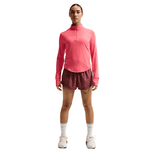 En person står og kigger fremad iført en Nike Swift UV Half Zip Running Top til kvinder i Sea Coral/Reflective Silver, parret med rødbrune shorts, hvide strømper og hvide sneakers på en almindelig hvid baggrund.