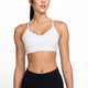 En kvinde står model for Nike Padded Adjustable Sports Bra til kvinder i White/Stone Mauve sammen med sorte leggings. Hun står på en almindelig hvid baggrund og kigger fremad med let drejet hoved og små stjernetatoveringer på armene.