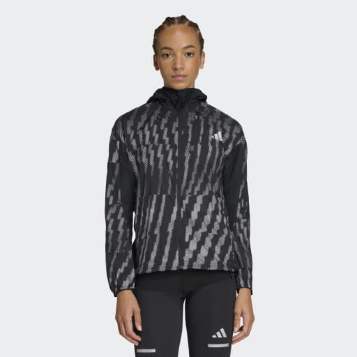 En person iført Adidas Adi365 Jacket, Dame, i Black/Refsil med gennemgående lynlås foran og sorte atletiske leggings står på en almindelig lysegrå baggrund.