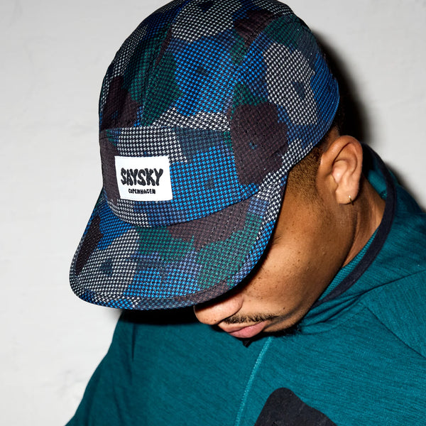 En person iført en blågrøn hættetrøje og Saysky Flower Combat Cap 103 (Unisex, Blue Aop) med et hvidt SKYSKY COPENHAGEN-mærke kigger ned, mens ansigtet er delvist skjult.