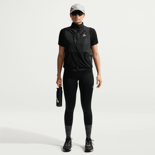 En person ser frem for sig i sorte Nike ACG High-Waisted 7/8 Running Tights (Dame, Black/Summit White), kortærmet vest med lynlås, sneakers, grå kasket og solbriller. De holder en vandflaske med en almindelig hvid baggrund.