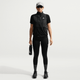 En person ser frem for sig i sorte Nike ACG High-Waisted 7/8 Running Tights (Dame, Black/Summit White), kortærmet vest med lynlås, sneakers, grå kasket og solbriller. De holder en vandflaske med en almindelig hvid baggrund.