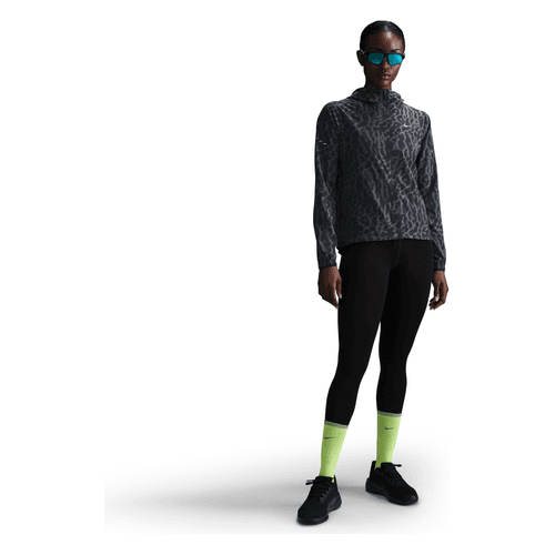 En person står på en hvid baggrund iført en mønstret mørk sweatshirt, sorte leggings, lysegrønne sokker, sorte sportssko, solbriller og Nike Dri-FIT løbejakke for ekstra synlighed.