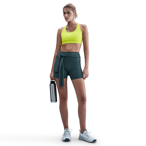 En kvinde ser frem for sig i en Nike Swoosh Medium Sports Bra, Dame, Cyber/White, sammen med mørke shorts og hvide sneakers. Hun holder en vandflaske af metal og har håret sat op i en hestehale. Baggrunden er hvid.