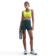 En kvinde ser frem for sig i en Nike Swoosh Medium Sports Bra, Dame, Cyber/White, sammen med mørke shorts og hvide sneakers. Hun holder en vandflaske af metal og har håret sat op i en hestehale. Baggrunden er hvid.