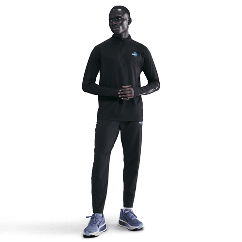 En person står på en hvid baggrund iført Nike Stride Dri-FIT 1/2-Zip Running Top, Herre, i sort, sammen med sorte atletiske bukser og blå atletiske sko.