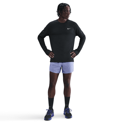 En person står med hænderne på hofterne iført Nike Dri-FIT Running Waffle Crew til mænd i Black/Reflective Silver, lyslilla shorts, sorte sokker og sorte Nike-løbesko med blå snørebånd på en almindelig hvid baggrund.