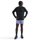 En person står med hænderne på hofterne iført Nike Dri-FIT Running Waffle Crew til mænd i Black/Reflective Silver, lyslilla shorts, sorte sokker og sorte Nike-løbesko med blå snørebånd på en almindelig hvid baggrund.