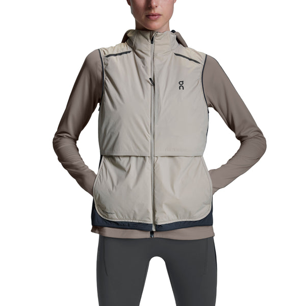 En kvinde står med hænderne på hofterne mod en hvid baggrund, iført en On Weather Vest Dame i Desert/Eclipse over en langærmet taupe top og sorte leggings.